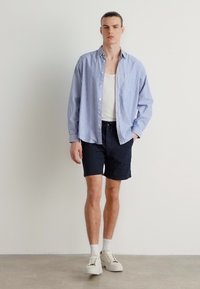 Camisa de botão às riscas, azul e branca, vestida sobre uma t-shirt branca, combinada com calções escuros e ténis brancos, de pé contra um fundo neutro.