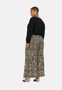 Žena s kratkom kosom nosi crnu majicu dugih rukava, maksi suknju sa zebrastim uzorkom, zlatne kružne naušnice i smeđe sandale s otvorenom petom, stoji bočno.