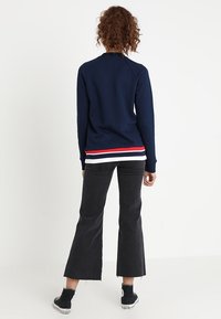 Námořnický pulovr s kulatým výstřihem a dlouhými rukávy, zdobený červenými a bílými pruhy na lemu. V kombinaci s rozšířenými černými džíny a teniskami.