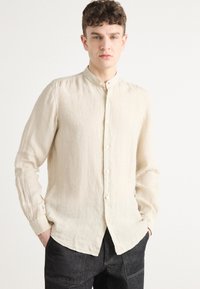 Camicia button-up beige in lino con colletto alla cinese, dotata di maniche lunghe e orlo dritto, abbinata a jeans scuri.
