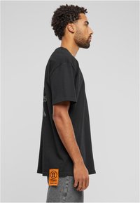 Juodas medvilnės marškinėliai trumpomis rankovėmis, laisvas kirpimas, su grafiniu dizainu ir oranžine etikete apatiniame krašte.