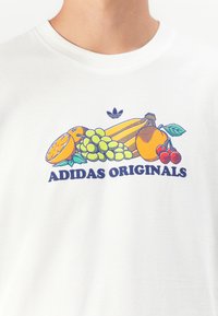 T-shirt bianca in cotone con una grafica di frutta colorata (arancia, uva, banana, ciliegia) e testo "ADIDAS ORIGINALS" in blu.