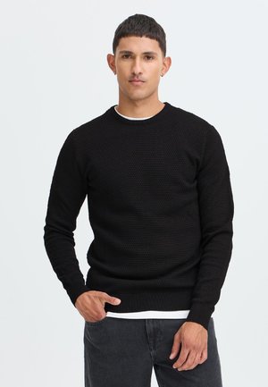 Homme aux cheveux courts et foncés portant un pull noir texturé par-dessus une chemise blanche et un jean foncé, debout devant un fond clair uni.