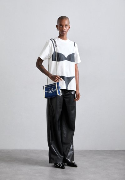 Tricou alb cu model abstract negru, pantaloni negri din piele cu croială largă și o geantă de umăr mică, albastră, cu text alb și curea din lanț argintie.