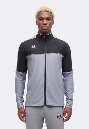 Joven con cabello corto rizado y rubio que lleva una chaqueta deportiva con cremallera gris y negra de Under Armour y pantalones a juego, de pie frente a un fondo liso.