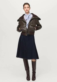 Femme portant une veste en cuir marron foncé par-dessus un haut bleu clair noué, une jupe évasée bleu marine et des bottes en cuir marron jusqu'aux genoux, se tenant avec les mains dans les poches.