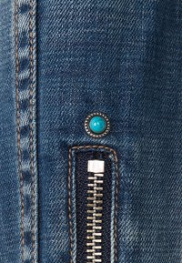 Denimkangas tummansinisellä värillä ja näkyvillä ompeleilla. Varustettu hopeisella vetoketjulla ja turkoosilla koristepainikkeella. Lähikuva.