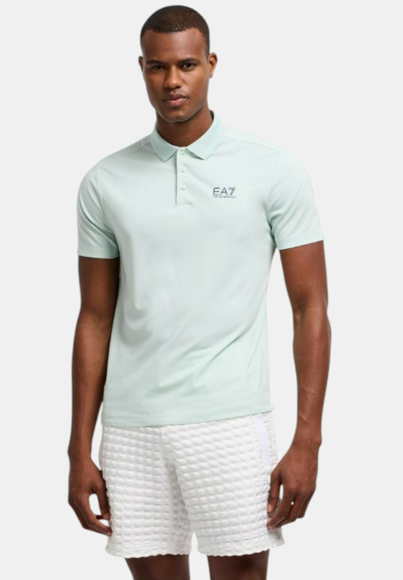 Polo de color verde pálido con tres botones y logo de EA7, combinado con pantalones cortos blancos texturizados con un patrón de cuadros.