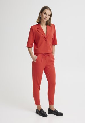 Γυναίκα που στέκεται με τα χέρια στις τσέπες, φορώντας κόκκινο σακάκι με κοντά μανίκια και ταιριαστό κόκκινο παντελόνι σε στενή γραμμή με μαύρα μοκασίνια.