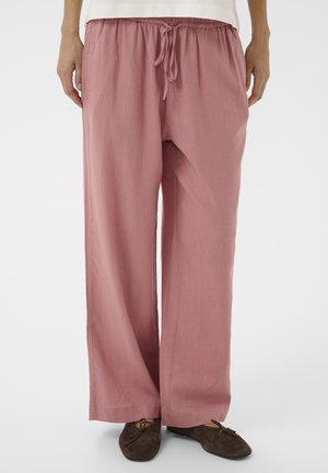Persona con pantalones rosa sueltos con cordón y zapatos planos marrones, de pie con las manos relajadas a los lados.
