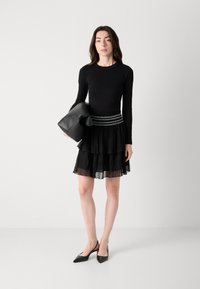 Svart langermet topp kombinert med en svart flosset mini nederdel med lagdeling og strukturert liv. Modellen holder en svart clutch og har på seg svarte hæler.