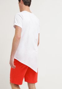 Camiseta blanca de cuello redondo con mangas enrolladas, combinada con pantalones cortos rojos. La camiseta tiene un dobladillo trasero más largo, lo que proporciona una silueta distintiva.