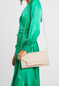 Béžová psaníčko s texturovaným povrchem a zlatým kováním, zobrazené na pozadí zelených saténových šatů s dlouhými rukávy a zavinovacím designem.