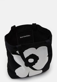 Black canvas tote bag with a large white floral design and a black center. Double shoulder straps and a branded label inside.