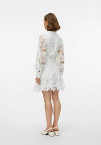 Weißes Spitzenkleid mit floralen Mustern, langen Ärmeln, tailliertem Bund und schwingendem Saum. Verfügt über eine zarte Textur und Transparenz.