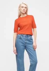 Νεαρή γυναίκα με ίσια ξανθά μαλλιά, ντυμένη με πορτοκαλί πουλόβερ με κοντά μανίκια και ψηλόμεσο μπλε τζιν, στέκεται μπροστά σε λευκό φόντο.