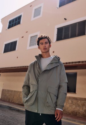 Młody mężczyzna z kręconymi włosami, ubrany w jasnoszarą kurtkę i białą koszulę, stoi na ulicy przed beżowym budynkiem z oknami.