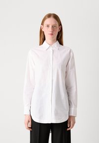 Camicia bianca a maniche lunghe con colletto, caratterizzata da una texture liscia. Indossata con pantaloni neri, presenta dettagli di bottoni visibili e un orlo laterale slit.