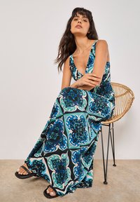 Maxi-jurk met een V-hals, met een kleurrijke bloemenprint in blauw, groen en zwart op een crèmekleurige achtergrond. Gecombineerd met platte zwarte sandalen.