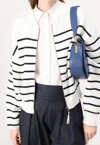 Biały kardigan na zamek z czarnymi paskami, o prążkowanej fakturze i kołnierzem typu szal, noszony na białej koszuli z guzikami, w połączeniu z ciemnymi spodniami.