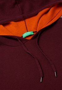 Sudadera burdeos con un interior naranja. Cuenta con cordones ajustables con puntas metálicas y una etiqueta verde de "United Colors of Benetton".