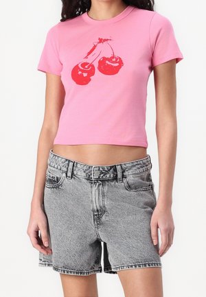 Mujer con una camiseta rosa corta con estampado de cerezas rojas combinada con pantalones cortos vaqueros grises de cintura alta, de pie frente a un fondo liso.