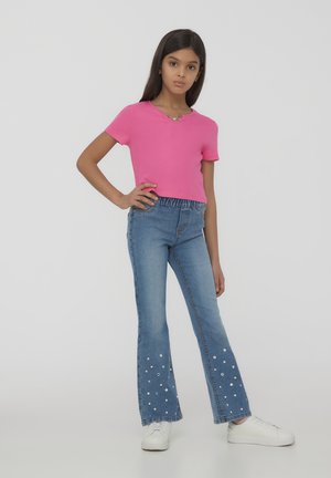 Top rosa a maniche corte con scollo a V abbinato a jeans a zampa d'elefante blu con decorazioni bianche all'orlo, indossato con sneakers bianche.