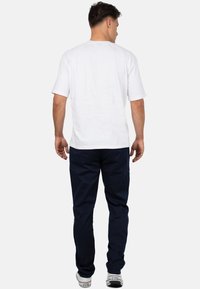 T-shirt bianca a maniche corte con colletto rotondo, abbinata a pantaloni blu navy e sneaker bianche, vista da dietro.
