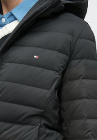 Fekete, steppelt mintás pufi dzseki, sima tapintású, cipzáras, a mellrészen kis logóval, amely piros, fehér és kék színű díszítéseket tartalmaz.