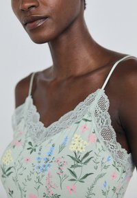 Groene bloemige camisole met kantafwerking, met roze, blauwe en gele bloemenpatronen. Dunne bandjes en een zachte stoftextuur.