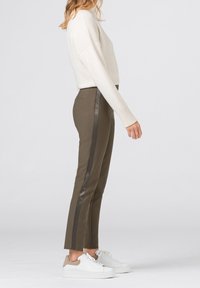 Pantalons ajustés olive avec des rayures noires sur les côtés, associés à un pull crème et des baskets blanches, présentés de profil.
