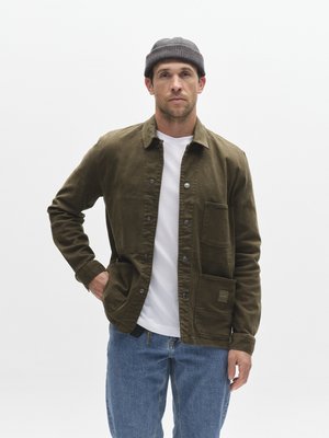 Oliven grønn bomullsjakke med knappelukking, to store framlommer og en brystlomme. Kombinert med en hvit T-skjorte og jeans.