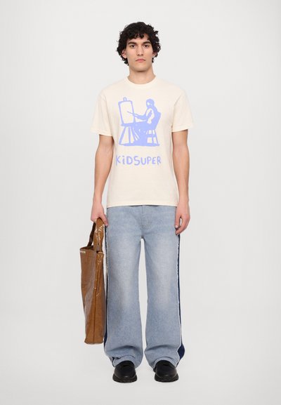 Cremefarbener Baumwoll-T-Shirt mit blauem Grafikdesign, kombiniert mit hellblauen weitgeschnittenen Jeans mit seitlichen Streifen und einer braunen Tragetasche.