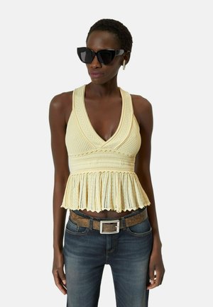 Vrouw met zwarte oversized zonnebril, mouwloos crème gebreid topje met een gestructureerd patroon en een gerimpelde zoom, donkere spijkerbroek met bruine riem.