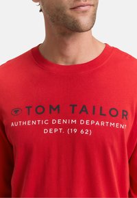 Rotes Langarm-T-Shirt mit dem Text "TOM TAILOR" und "AUTHENTIC DENIM DEPARTMENT" in Schwarz. Weicher Baumwollstoff mit einem klassischen Rundhalsausschnitt.