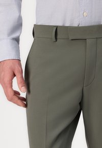 Olivgrön skräddarsydda byxor med en mjuk textur, platt fram, och bälteshällor, matchade med en ljusblå långärmad skjorta.