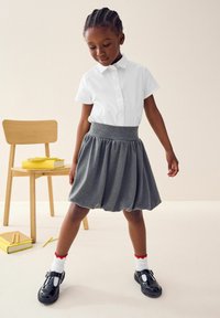 Chemise blanche à manches courtes avec col, jupe bulle grise avec taille élastique, chaussures en cuir verni noir et chaussettes blanches avec bord rouge.