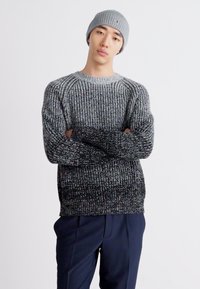 Joven con un gorro de punto gris, suéter grueso de punto degradado en negro y gris, y pantalones oscuros azules, de pie con los brazos cruzados.