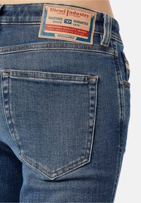 Vista trasera de una persona que lleva puestos unos vaqueros de denim azul con un gran bolsillo trasero y una etiqueta de cuero marrón de la marca Diesel Industry en la parte superior.