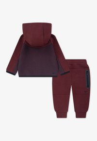 Bordový Nike joggingový set pozostávajúci z mikiny s kapucňou a zodpovedajúcich nohavíc. Hladká tkanina s vreckom na zips na nohaviciach.