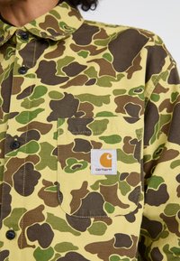 Koszulka bawełniana w żółtym kamuflażu z zielonymi i brązowymi akcentami. Posiada czarne guziki, kieszeń zapinaną na guzik oraz metkę z logo Carhartt.