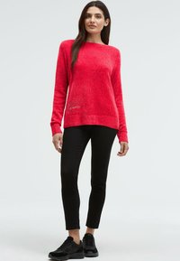 Eine Frau steht in einem roten Langarmpullover, schwarzen Slim-Hosen und schwarzen Sneakers vor einem schlichten hellen Hintergrund.