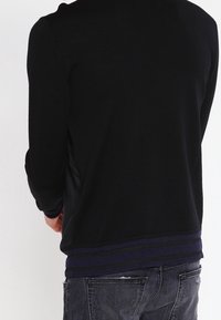 Black knit sweater with long sleeves and a ribbed hem featuring navy stripe accents. Fabric is smooth with a fitted design.
