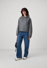 Grå, kort jakke med hette og elastiske mansjetter, kombinert med blå vid-benede jeans og hvite sneakers, supplert med en liten svart veske.