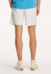 Hvide bomuldsbukser med en baglomme og lige snit, parret med beige høj-top sneakers med røde accenter og en logo patch.