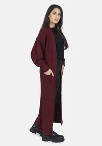 Cardigan lungo bordeaux in tessuto texturizzato, con apertura frontale e tasche laterali. Abbinato a un top nero e pantaloni neri, con stivali neri robusti.