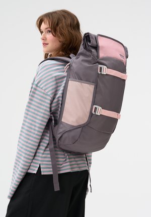 Junge Frau mit einem großen grauen und pinken Rucksack, gekleidet in ein gestreiftes Langarmshirt und schwarze Hose, die zur Seite schaut.