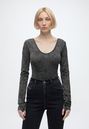 Lyshåret kvinde med kort hår iført en grå marmorprintet langærmet top og høj talje sorte jeans, stående mod hvid baggrund.