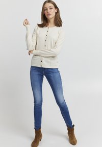 Cardigan crem cu tricot ușor, guler rotund, închidere cu nasturi în față, nasturi gri și mâneci lungi, asortat cu blugi skinny albaștri.