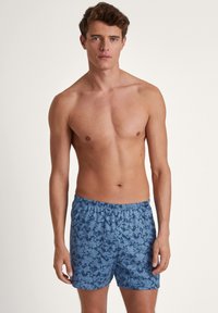 Blauwe gestreepte zwembroek met een elastische tailleband, met een bladmotief in verschillende tinten blauw. Lichtgewicht stof, lengte tot halverwege de dij.